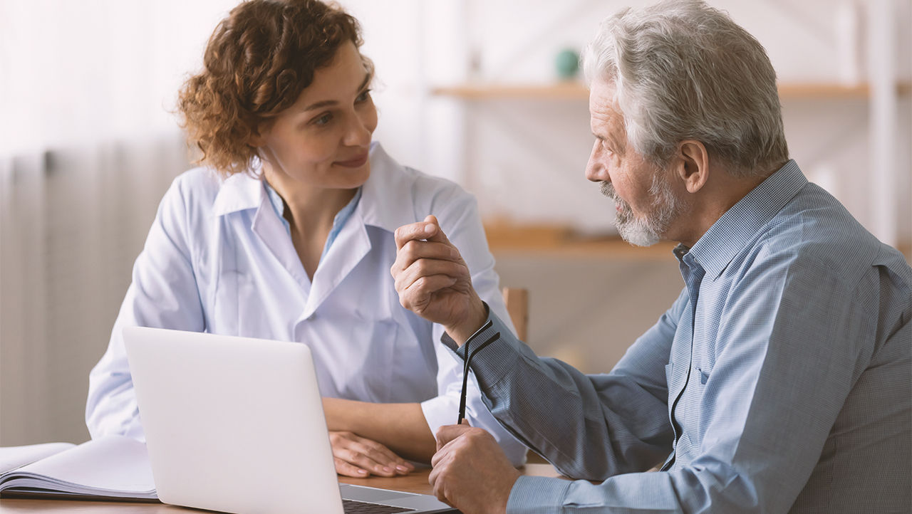 A physician reassuring an elderly male patient who is sitting across a table from her.