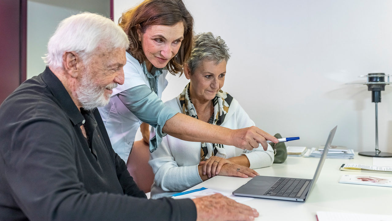 An older man and woman look across a table at a tablet held by a woman doctor in a white coat.