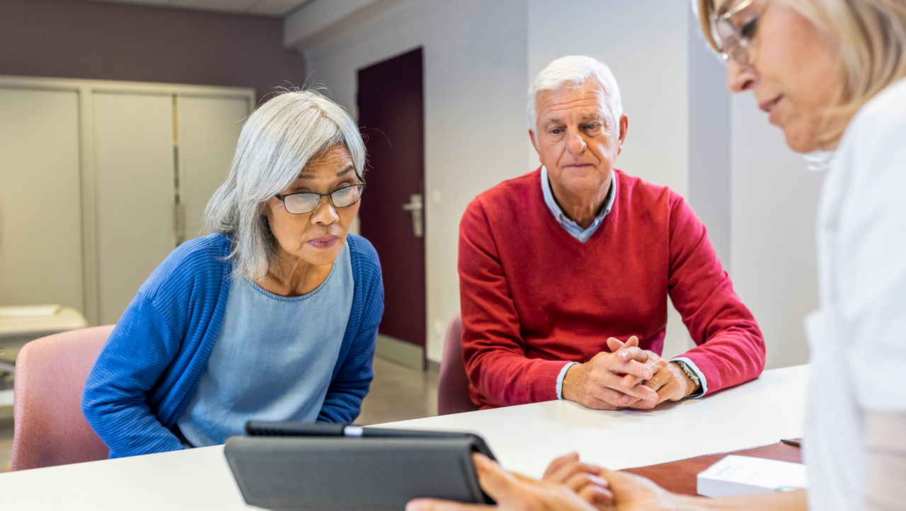 An older man and woman look across a table at a tablet held by a woman doctor in a white coat.