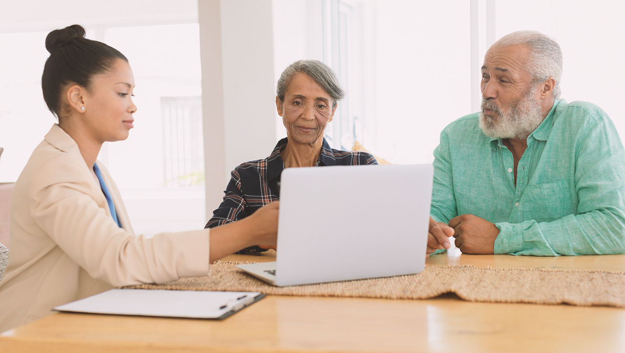 An older man and woman look across a table at a tablet held by a woman doctor in a white coat.