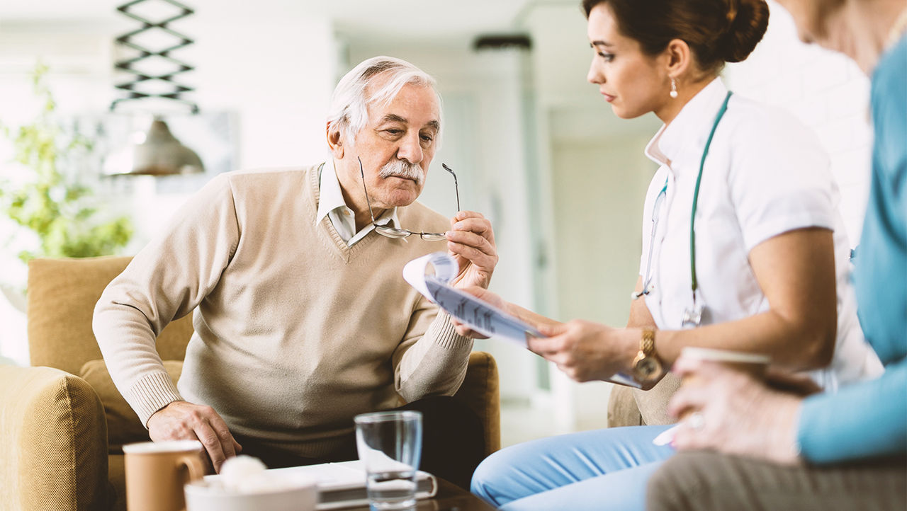 An older man and woman look across a table at a tablet held by a woman doctor in a white coat.