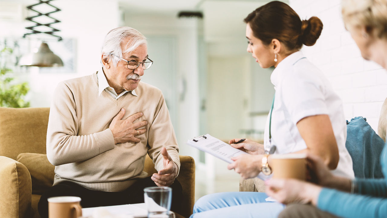 A doctor in a white coat is sitting behind a desk with a notepad. She is explaining the male sling system to her elderly patients.